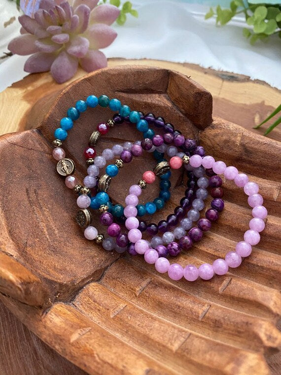 Stackable gemstone beaded bracelets in purple, lavender, and teal displayed on a rustic wood tray with succulents, featuring gold accent beads and a saint medal charm