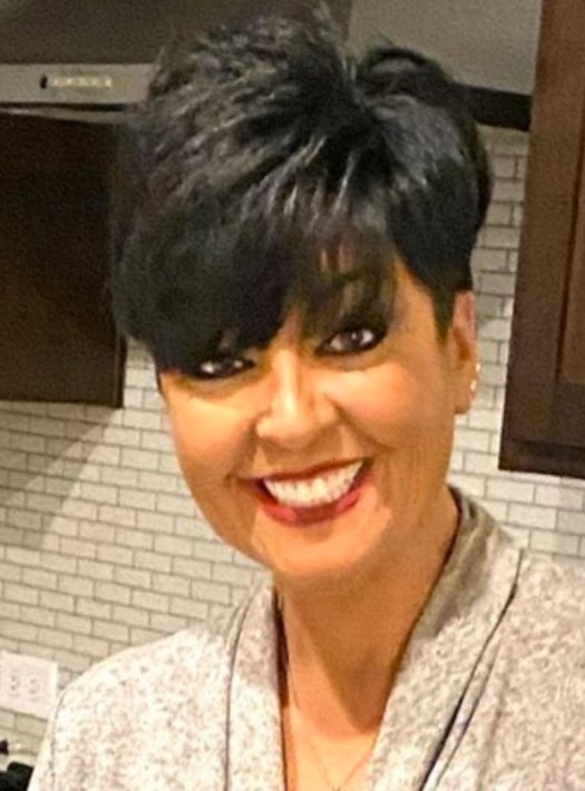 Professional headshot of Maria Oren, smiling woman with short dark hair and red lipstick, wearing a grey top in front of a white brick backsplash