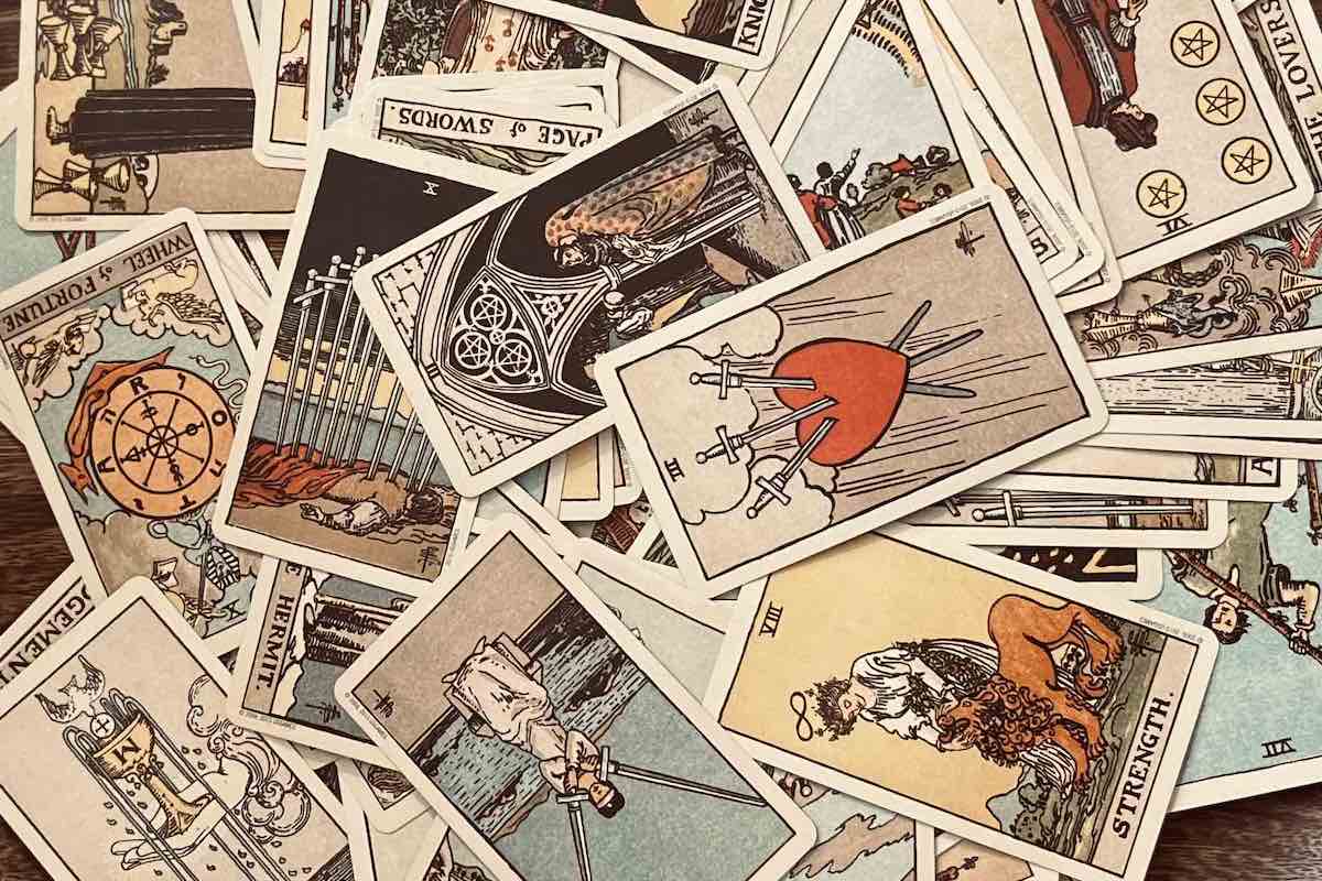 A scattered pile of Rider-Waite tarot cards spread across a wooden surface, with visible cards including the Three of Swords, Wheel of Fortune, The Hermit, Strength, Page of Swords, and The Lovers