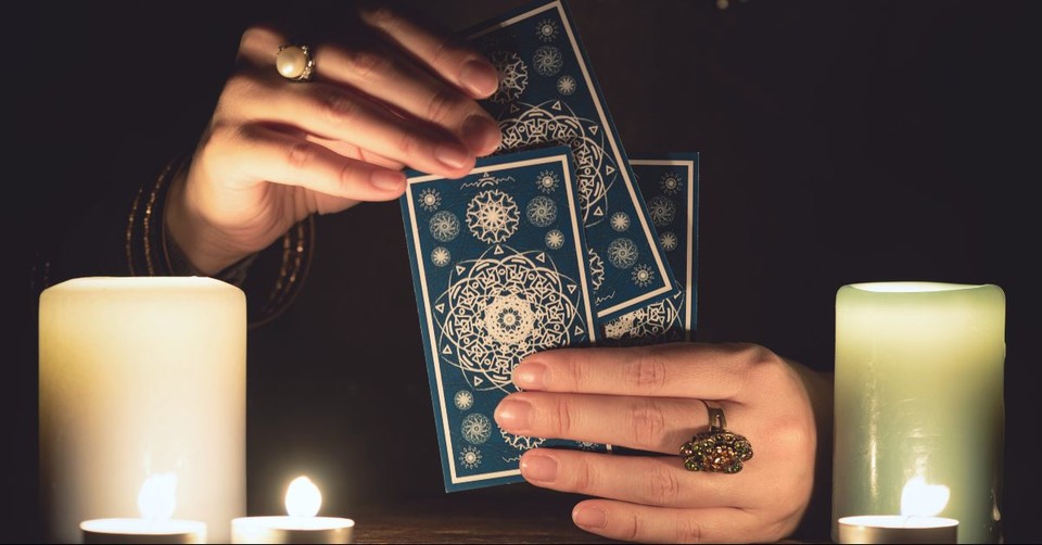 Close-up of a woman's ringed hands pulling tarot cards from a blue mandala-patterned deck by candlelight, with two large white pillar candles glowing on either side in a dark, atmospheric setting