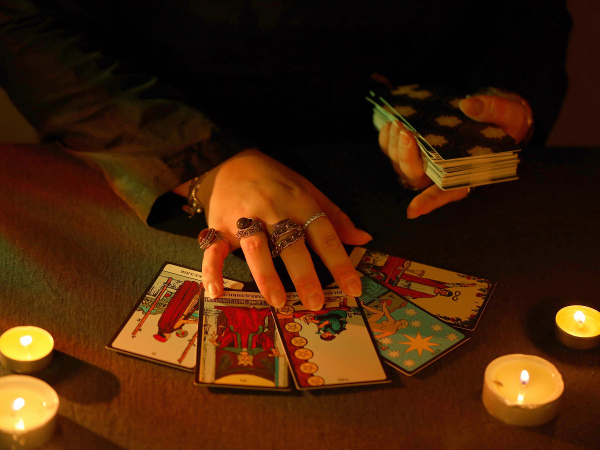 A tarot card reader with silver rings spreads Rider-Waite tarot cards across a dark table by candlelight, holding the remaining deck in one hand while surrounded by glowing tealight candles in a moody, mystical atmosphere