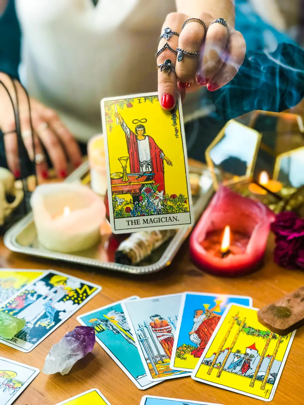 A bejeweled hand with silver rings and red nails holds up The Magician tarot card during a reading, surrounded by lit candles, an amethyst crystal, sage, and a spread of Rider-Waite tarot cards on a wooden table
